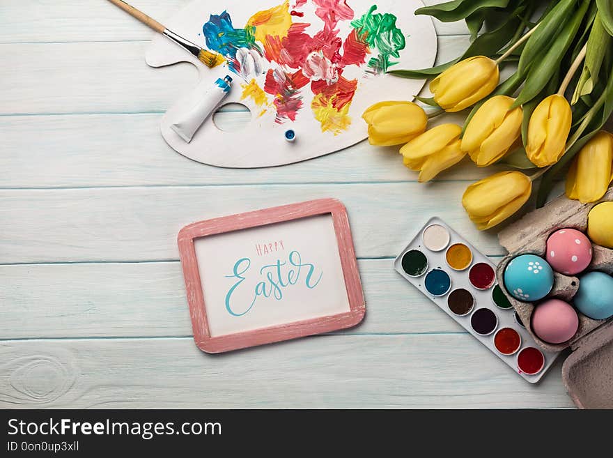 Colorful easter eggs, paint palette, brushes and yellow tulips on white wooden table. Top view with copy space