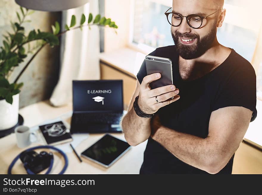 Portrait smiling handsome man reading text message, standing indoors. In background is laptop with inscription- e-learning on monitor.Businessman sends messages.Stylish brunette man uses mobile phone. Portrait smiling handsome man reading text message, standing indoors. In background is laptop with inscription- e-learning on monitor.Businessman sends messages.Stylish brunette man uses mobile phone