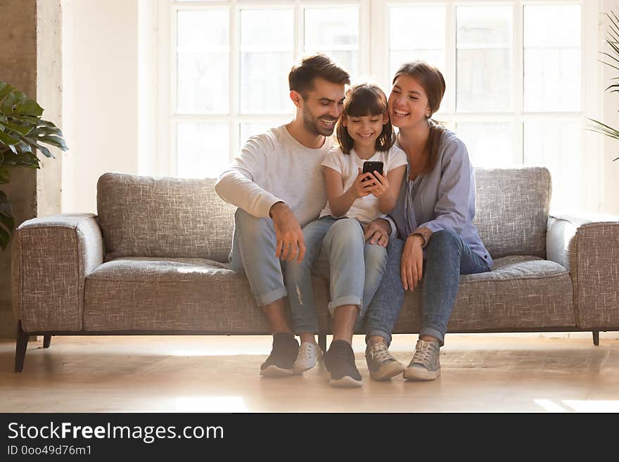 Happy family and kid daughter having fun with smartphone gadget at home, little child girl looking at phone play game using app with mom dad watching funny mobile video, making online call on couch. Happy family and kid daughter having fun with smartphone gadget at home, little child girl looking at phone play game using app with mom dad watching funny mobile video, making online call on couch