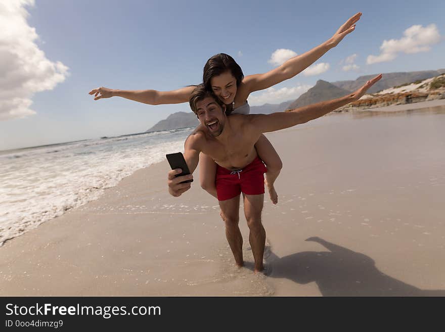 Front view of happy young couple piggyback with arms outstretched taking selfie with mobile phone at beach in the sunshine