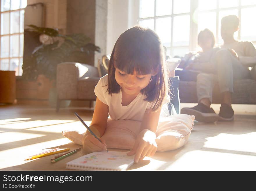 Cute happy little kid girl drawing with colored pencils lying on warm floor in light cozy living room, smiling preschool child daughter enjoy creative activity while parents relaxing on couch at home. Cute happy little kid girl drawing with colored pencils lying on warm floor in light cozy living room, smiling preschool child daughter enjoy creative activity while parents relaxing on couch at home