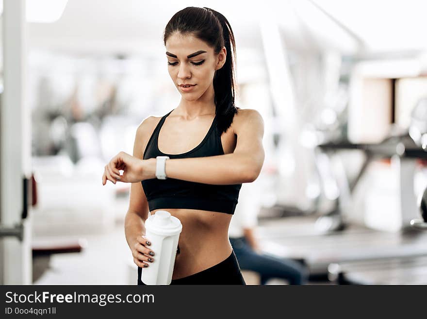 Beautiful slim girl dressed in black sportswear is standing in the gym and looking at her watch .