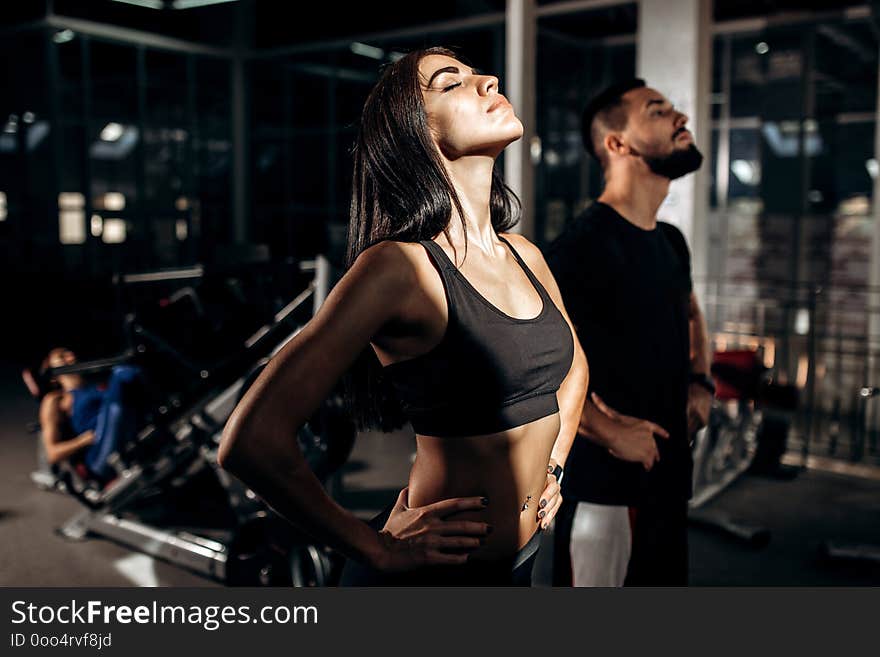 Slim dark-haired girl dressed in black sport clothes and fitness coach are doing warm up in the gym .