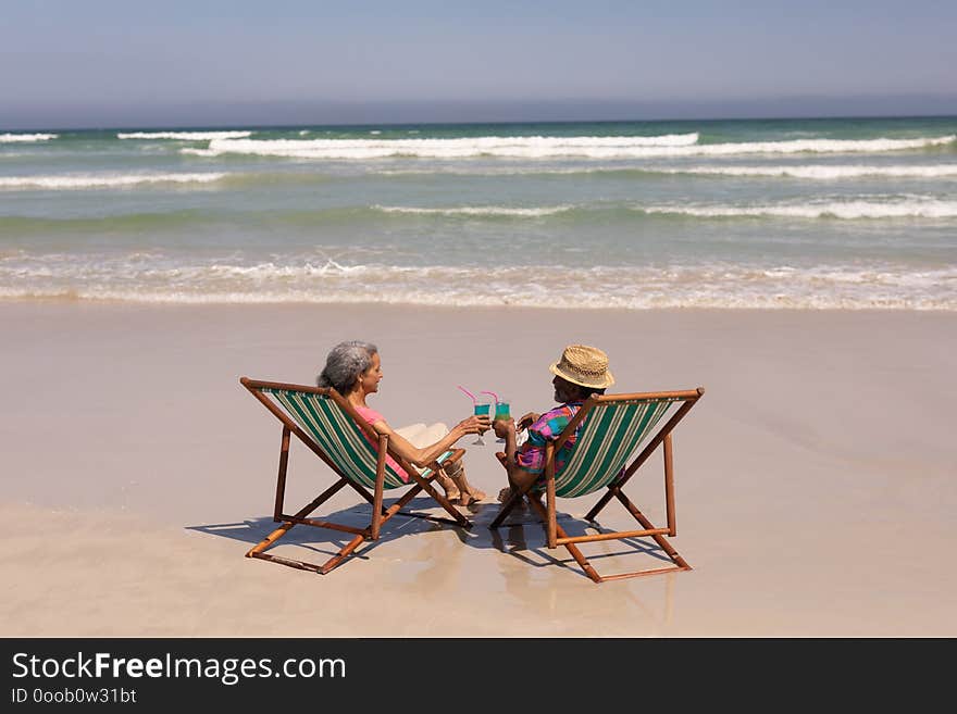 Rear view of senior couple relaxing on sun lounger and toasting cocktail glasses on beach in the sunshine