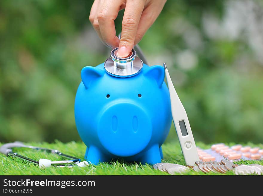 Woman using stethoscope to check blue piggy bank with thermometer and medicine on natural green background,Save money for Medical