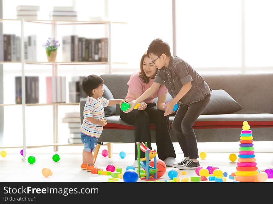 Asian family, happy time at home, young beautiful mother, taking care sons, little adorable toddler, standing, young boy wearing eyes glasses hold green ball, playing together in modern living room. Asian family, happy time at home, young beautiful mother, taking care sons, little adorable toddler, standing, young boy wearing eyes glasses hold green ball, playing together in modern living room