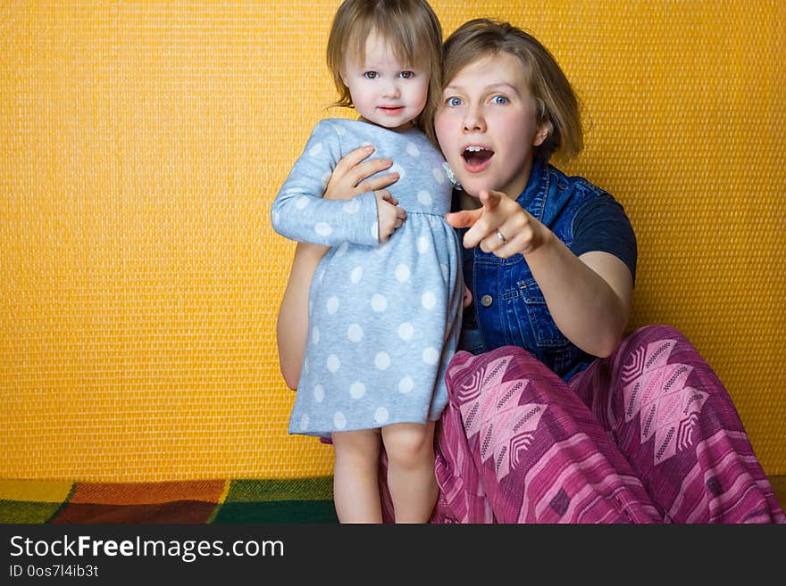 Portrait of mother hugging toddler daughter. pretty, amazed girl look to camera. woman, person point at surprise by finger. cute, adorable child loving her mommy. mom have blue eyes and blond hair.