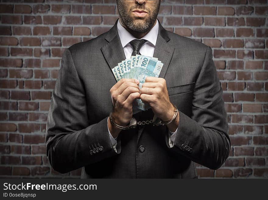 Brazilian man holding bills of money with a handcuff in a jail. concept of corruption, corrupt politicians, illegal businesses. brick background. Brazilian man holding bills of money with a handcuff in a jail. concept of corruption, corrupt politicians, illegal businesses. brick background