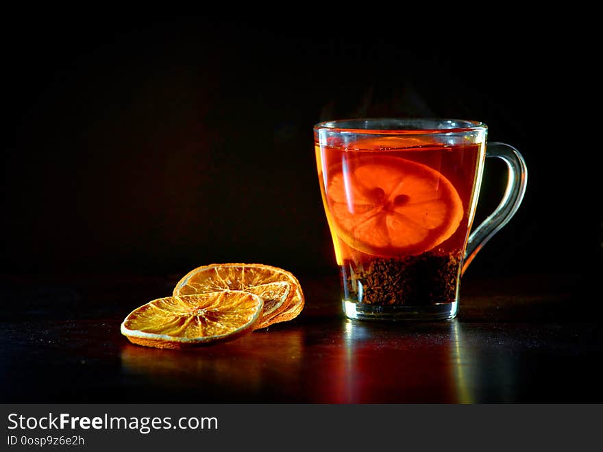 A cup of black tea and lemon, pieces of dried orange. Dark background, warm shades, steam