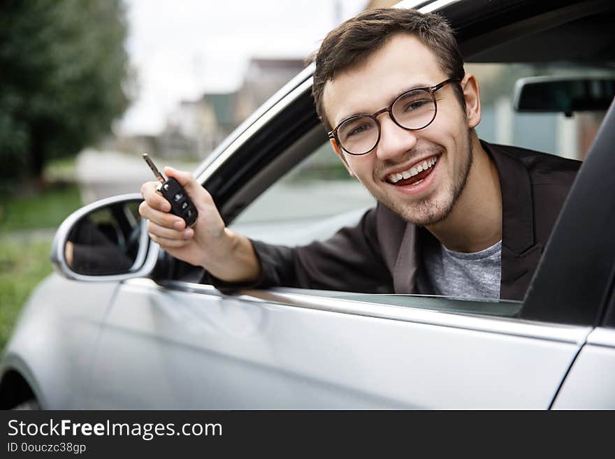 Satisfied young guy is peeking from the car window while looking at the camera. He is holding the keys at his right hand. Lottery winner concept.