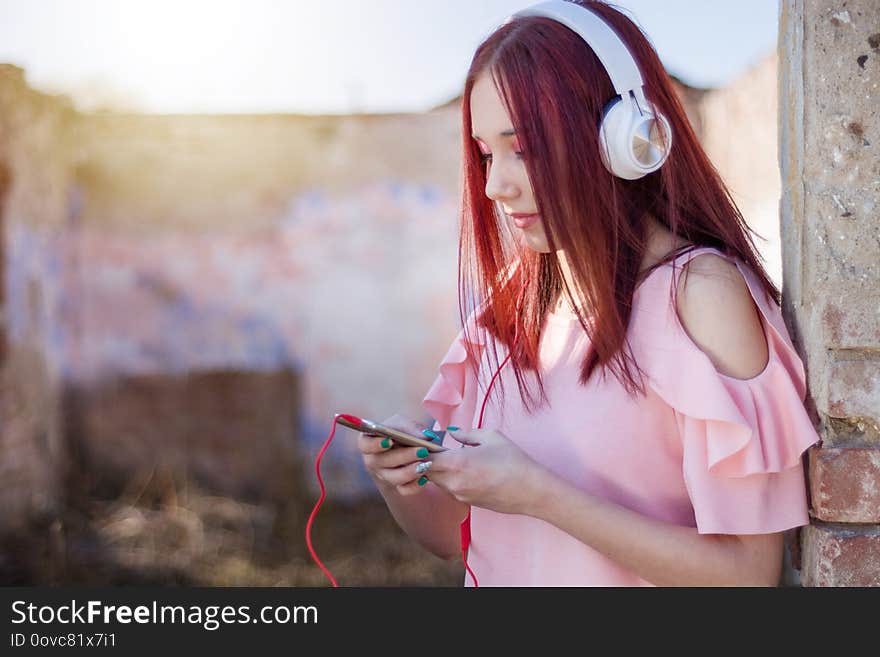 This captivating image portrays a young redhead teenager girl enjoying a moment of solitude and musical bliss against the backdrop of a mesmerizing sunset. Sitting alone on a retro ruins wall, she is fully immersed in her smartphone and wearing a headset, indulging in the pleasure of her favorite tunes. The close-up shot and selective focus draw attention to the teenager's expressive face, revealing her deep engagement with the music. Her eyes are closed, and a serene smile graces her lips, reflecting the joy and emotional connection she feels. The blurred background adds to the dreamlike ambiance, emphasizing the focus on her and the music she's listening to. The juxtaposition of the old ruins wall with the modern smartphone and headset symbolizes the blending of past and present, showcasing the universal and timeless nature of music's ability to transport us to another world. The warm tones of the sunset provide a soft, golden glow, creating a tranquil and magical atmosphere. This image captures the essence of solitude, self-expression, and the power of music to transport us. It resonates with teenagers and music enthusiasts who seek moments of introspection and escape. It is perfect for artistic projects, music-related promotions, social media campaigns, and any visual content that aims to evoke a sense of nostalgia, serenity, and the transformative power of music. This captivating image portrays a young redhead teenager girl enjoying a moment of solitude and musical bliss against the backdrop of a mesmerizing sunset. Sitting alone on a retro ruins wall, she is fully immersed in her smartphone and wearing a headset, indulging in the pleasure of her favorite tunes. The close-up shot and selective focus draw attention to the teenager's expressive face, revealing her deep engagement with the music. Her eyes are closed, and a serene smile graces her lips, reflecting the joy and emotional connection she feels. The blurred background adds to the dreamlike ambiance, emphasizing the focus on her and the music she's listening to. The juxtaposition of the old ruins wall with the modern smartphone and headset symbolizes the blending of past and present, showcasing the universal and timeless nature of music's ability to transport us to another world. The warm tones of the sunset provide a soft, golden glow, creating a tranquil and magical atmosphere. This image captures the essence of solitude, self-expression, and the power of music to transport us. It resonates with teenagers and music enthusiasts who seek moments of introspection and escape. It is perfect for artistic projects, music-related promotions, social media campaigns, and any visual content that aims to evoke a sense of nostalgia, serenity, and the transformative power of music.