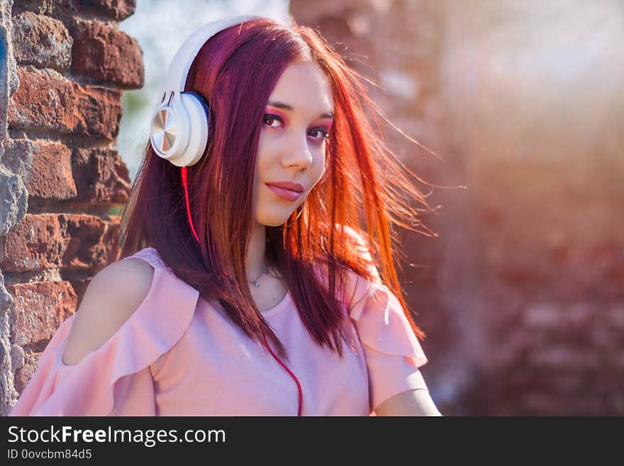 Gorgeous redheads lady listening music in headset on blurred background and red wall bricks in sunset. Charming girl with earphones enjoys with hair waving on wind. Close up, selective focus. Gorgeous redheads lady listening music in headset on blurred background and red wall bricks in sunset. Charming girl with earphones enjoys with hair waving on wind. Close up, selective focus