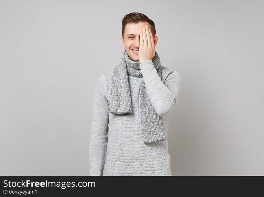 Smiling young man in gray sweater, scarf covering eyes with hand on grey wall background in studio. Healthy