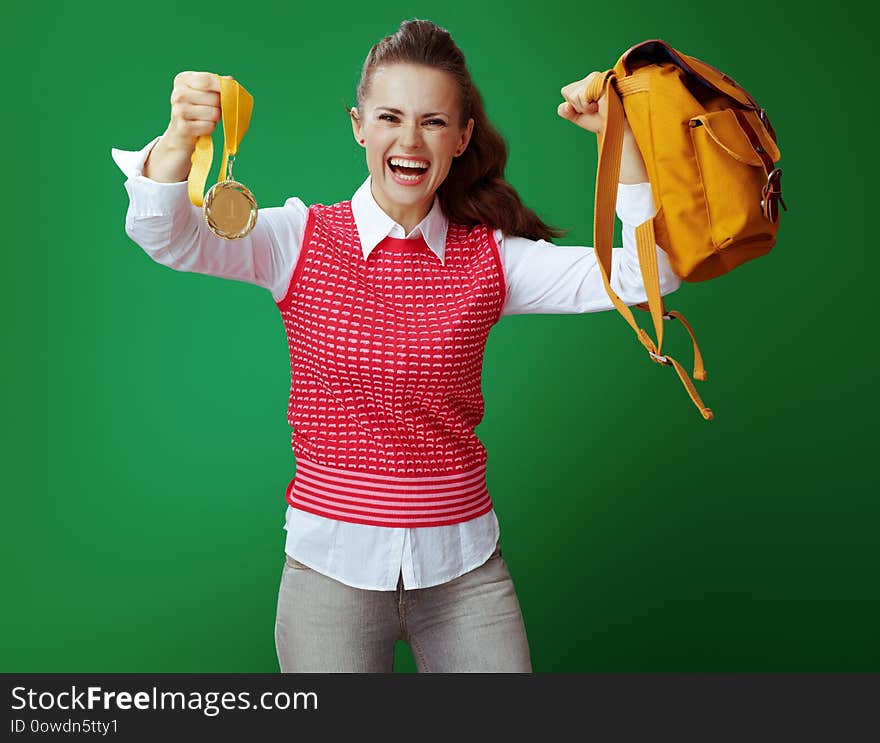 Smiling modern student woman in grey jeans and pink sleeveless shirt showing number one golden medal and biceps with a yellow backpack isolated on green background. Smiling modern student woman in grey jeans and pink sleeveless shirt showing number one golden medal and biceps with a yellow backpack isolated on green background