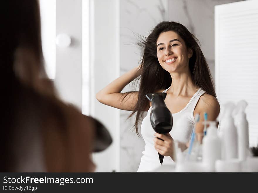 Smiling beautiful girl drying her hair in front of mirror in bathroom.