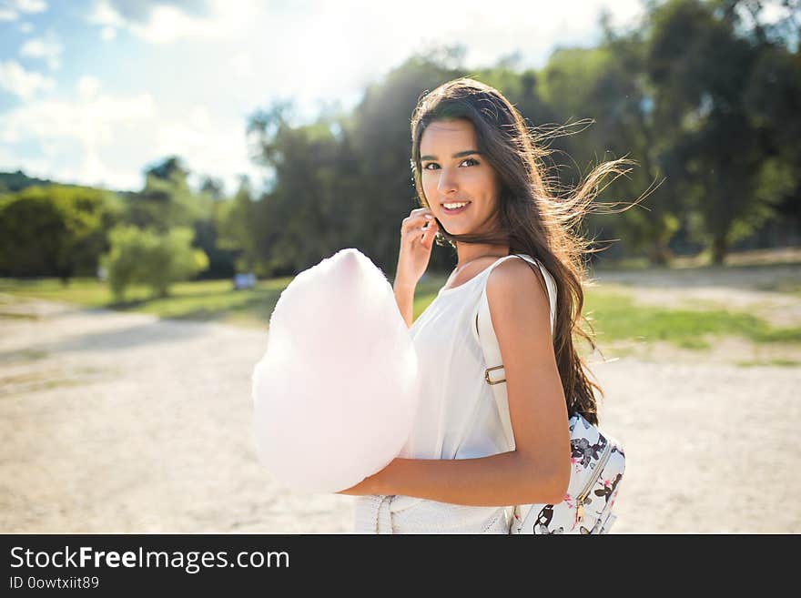 Beautiful brunette woman with cotton candy and windy hair, with backpack walking in the park and looking at the camera