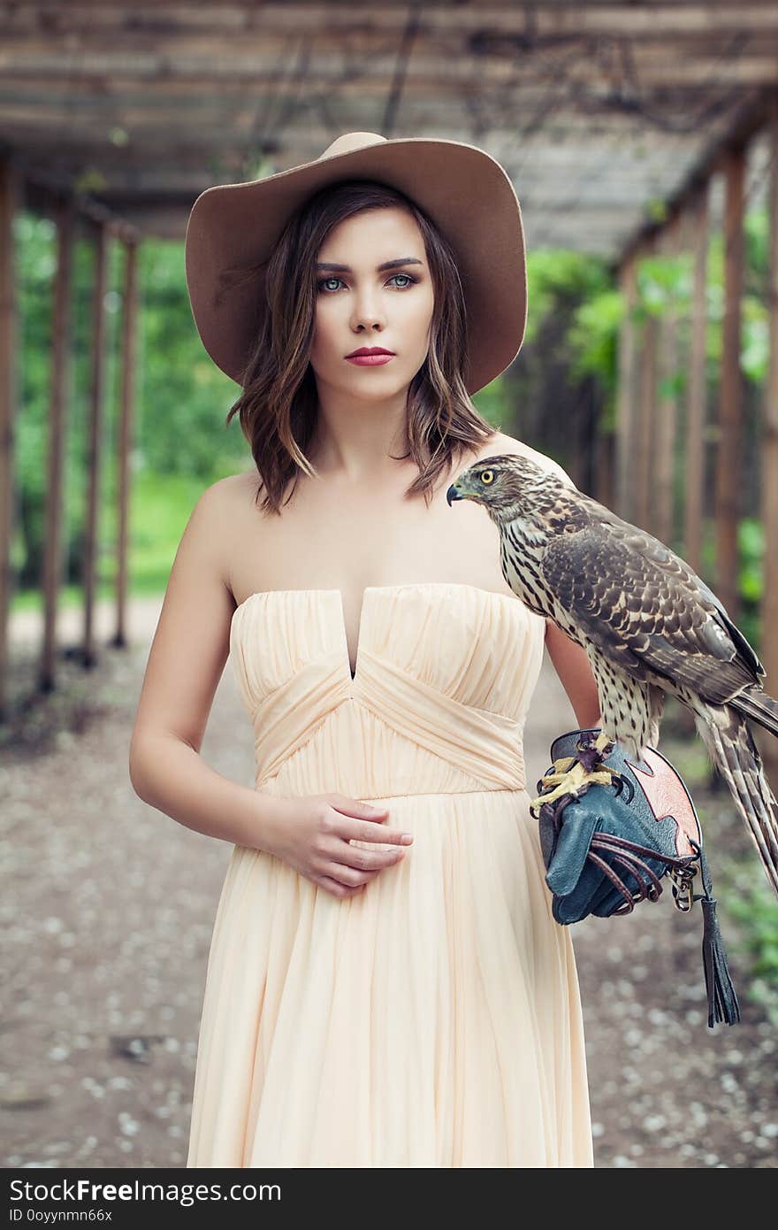 Sensual woman wearing peach color dress and brown hat with bird, outdoor fashion portrait.