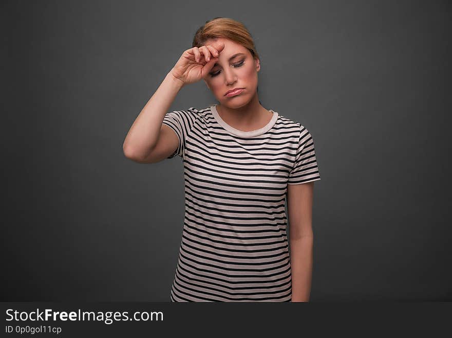 Tired woman on a gray background. The blonde holds her head with her eyes closed.