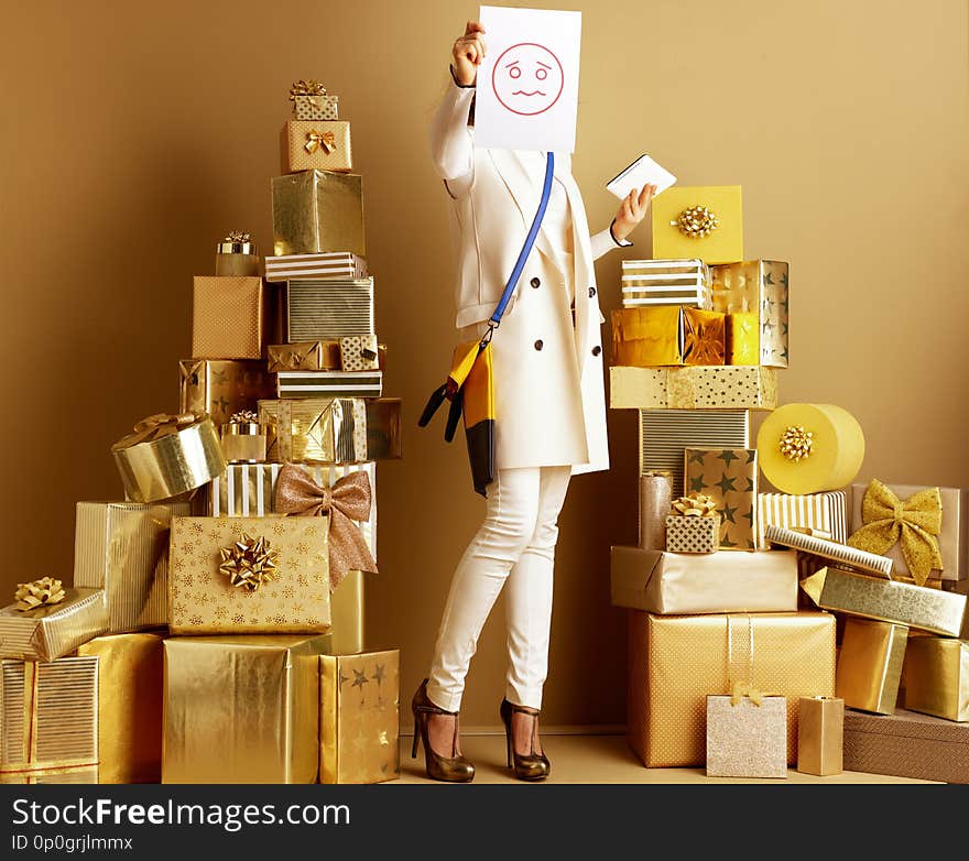 Stylish woman in white clothes with tablet PC and paper sheet with unhappy smile emoticon in the front of face symbolising bad review for product or retailer among 2 piles of golden gifts. Stylish woman in white clothes with tablet PC and paper sheet with unhappy smile emoticon in the front of face symbolising bad review for product or retailer among 2 piles of golden gifts