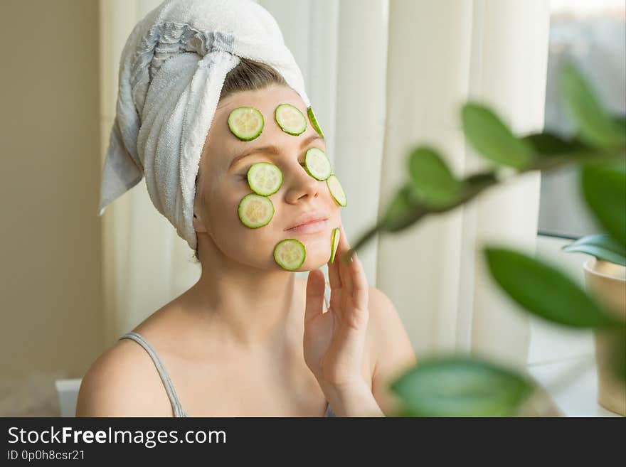 Young beautiful woman close-up at home near the window with natural homemade mask of cucumber on face, towel on head. Skin care, cosmetics, cosmetology, dermatology