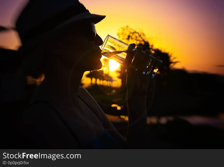 Woman with white hat and pink sunglasses with nice reflection of palm trees and sunset drinking fresh pure water outside on a sunny day from a glass