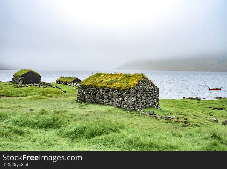 Beautiful fisherman rock house with green grass on the roof, sea side next to blue ocean with boat in the background foggy weather in Bour village, Faroe Islands, North Europe travel destination