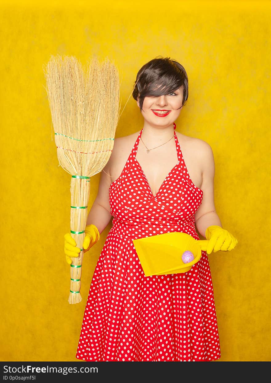 Cute charming emotional young woman in red retro polka dot dress stands in rubber gloves for cleaning with a broom in her hands for homework on a yellow solid background in the Studio alone, she scream and laugh