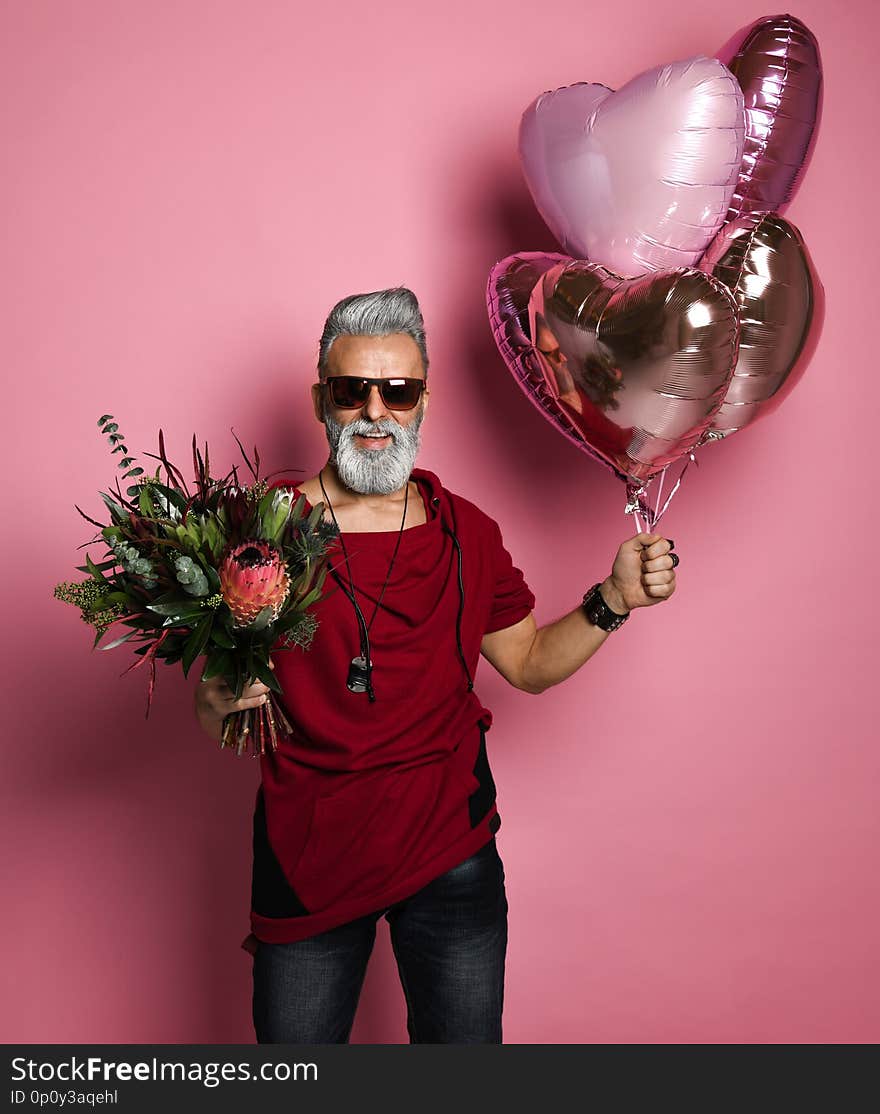 Stylish bearded middle-aged man with a modern hairstyle and fashionably dressed holding bunch of heart shaped balloons and a bouquet of flowers. Concept Valentine`s Day, Date. Stylish bearded middle-aged man with a modern hairstyle and fashionably dressed holding bunch of heart shaped balloons and a bouquet of flowers. Concept Valentine`s Day, Date