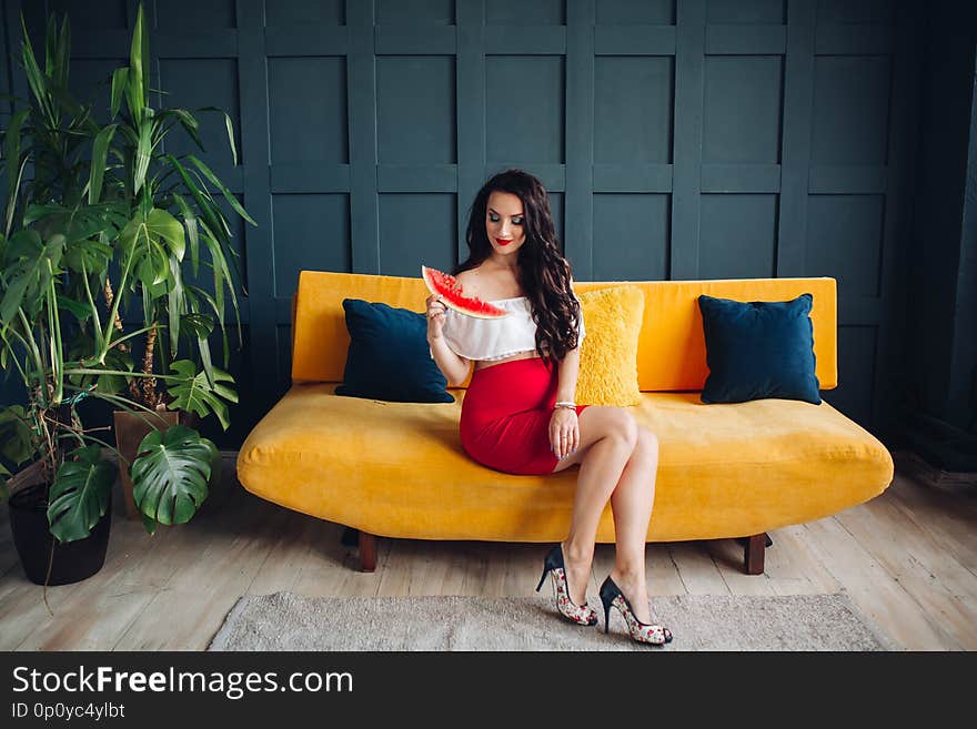 Stylish pregnant woman sitting on orange sofa, eating watermelon and posing. Attractive future mother in elegant clothes awaiting for little baby and relaxing. Concept of pregnancy and fashion. Stylish pregnant woman sitting on orange sofa, eating watermelon and posing. Attractive future mother in elegant clothes awaiting for little baby and relaxing. Concept of pregnancy and fashion.