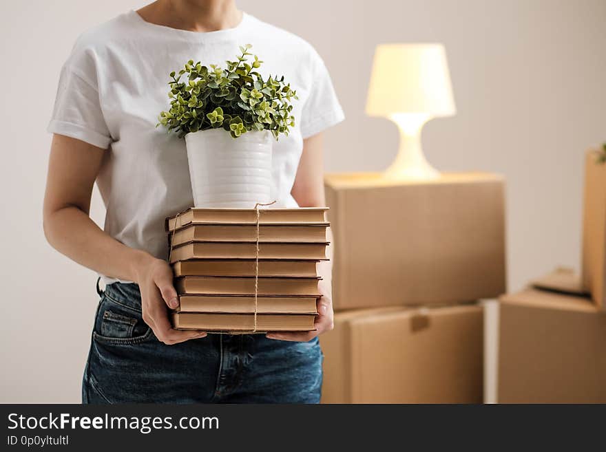 The concept of relocation and moving to a new home. Close-up, female hands hold a pile of books and a green plant in a pot. On the background of cardboard boxes and a table lamp.