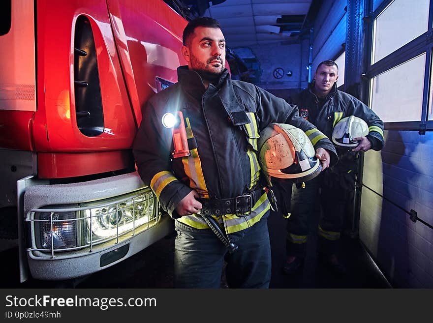 Two firemen wearing protective uniform standing next to a fire truck in a garage of a fire department.