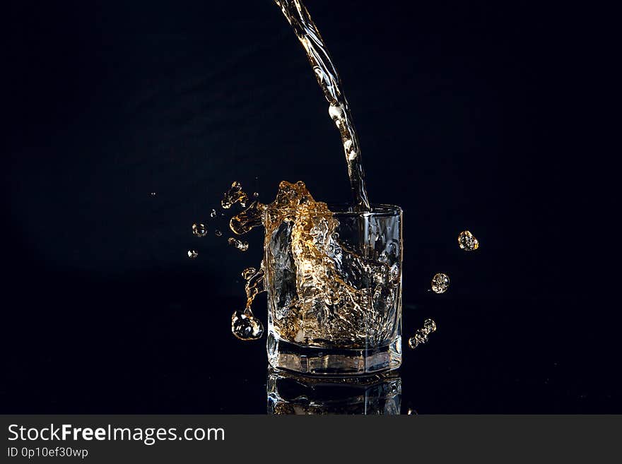 Whiskey pouring into glass with ice isolated on reflective black surface. Whiskey splash out of glass, many drops of beverage get out from glass. Whiskey pouring into glass with ice isolated on reflective black surface. Whiskey splash out of glass, many drops of beverage get out from glass