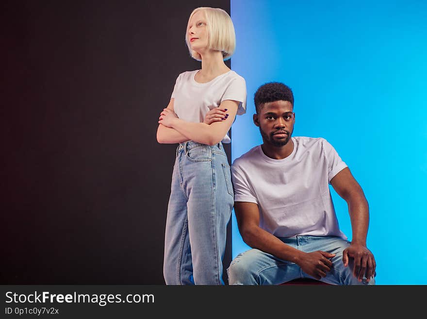 Attractive Afro men is sitting on the chair and looking at the camera, while his Caucasian albino girlfriend standing with crossed arms bear him, copy space. Attractive Afro men is sitting on the chair and looking at the camera, while his Caucasian albino girlfriend standing with crossed arms bear him, copy space