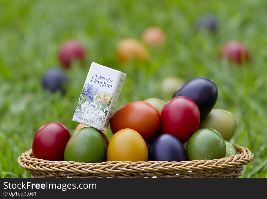 A basket of red, blue, green, orange and yellow painted eggs with a mini book, A gem of a daughter in some grass on a summer afternoon. A basket of red, blue, green, orange and yellow painted eggs with a mini book, A gem of a daughter in some grass on a summer afternoon