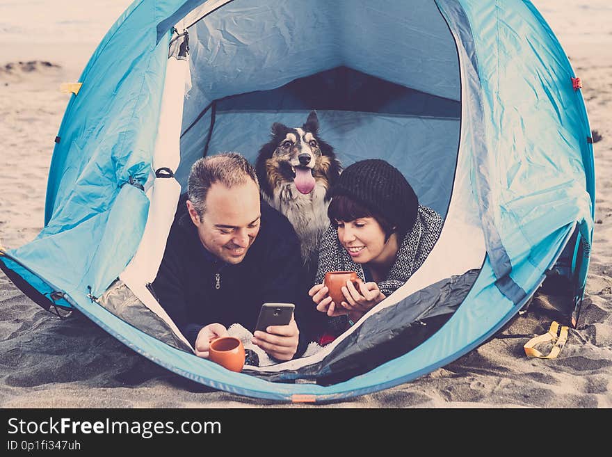 Couple looking at the smart phone and have fun inside a tent in free camping on the beach Dog border collie behind them looking at the camera. vintage colors and vacation family concept. alternative travel lifestyle. Couple looking at the smart phone and have fun inside a tent in free camping on the beach Dog border collie behind them looking at the camera. vintage colors and vacation family concept. alternative travel lifestyle