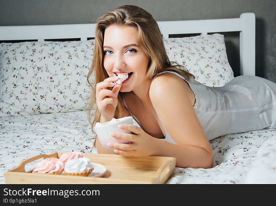 Young blonde with cupcakes eating marshmallow lying in bed