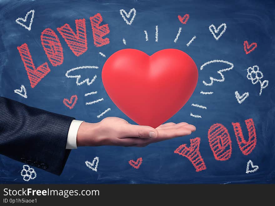 Male hand holding red heart with LOVE YOU chalk sign and heart drawings on blue background