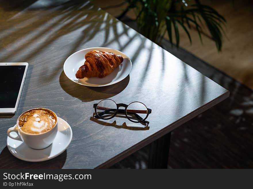 Close up of coffee cup, croissant, glasses and pad with blank screen