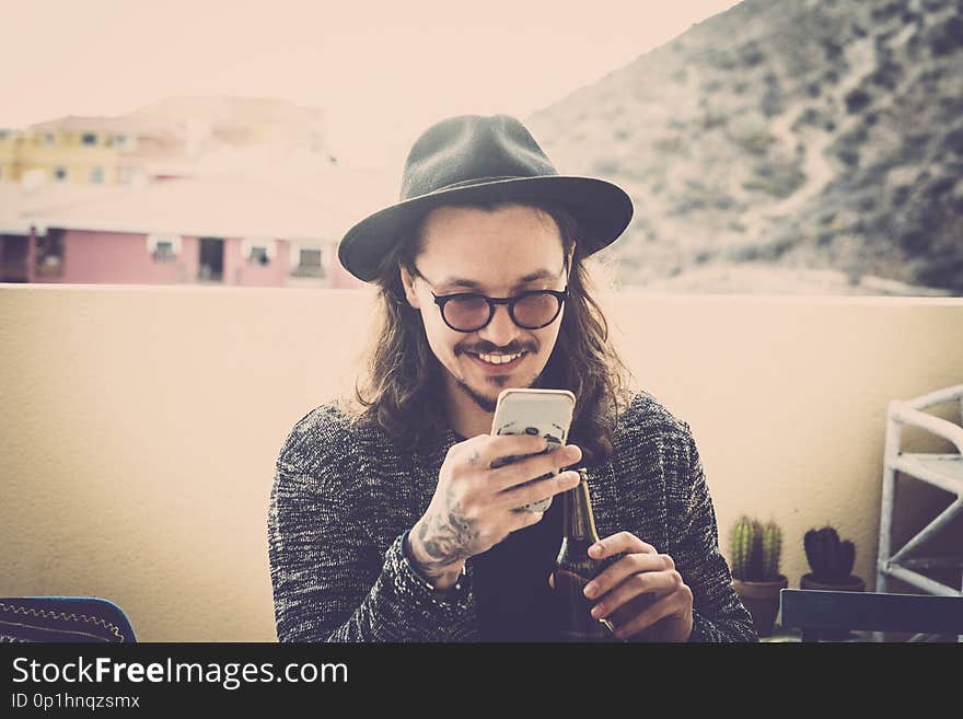 Long hair nice young man read and check the phone on the roofteop terrace outdoor. black hat and eyeglasses and tattoo on hand. fashion model sith nice smile. modern hipster concept. Long hair nice young man read and check the phone on the roofteop terrace outdoor. black hat and eyeglasses and tattoo on hand. fashion model sith nice smile. modern hipster concept