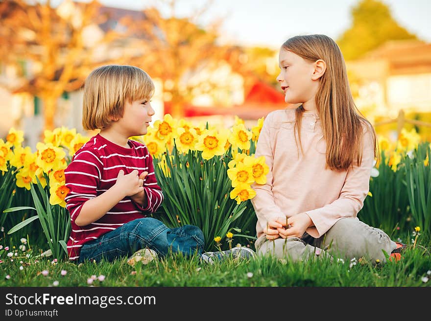 Two cute kids, little boy and his big sister, playing in the park between yellow daffodils flowers in sunlight