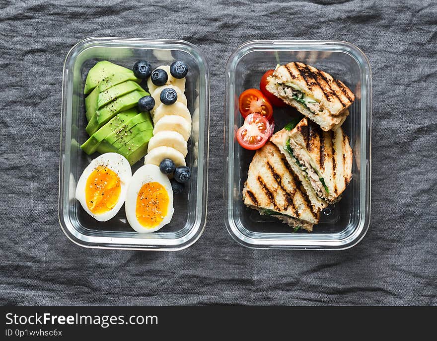 Two healthy office lunch box with sweet and savoury food. Boiled egg, avocado, tuna spinach cheese sandwiches and fruit on grey background, top view. Two healthy office lunch box with sweet and savoury food. Boiled egg, avocado, tuna spinach cheese sandwiches and fruit on grey background, top view