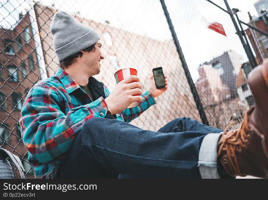 Young trendy man sitting on the street ewalk resting with cup of hot coffee and using smartphone for video link. Casual man calling on video link his friends. Young trendy man sitting on the street ewalk resting with cup of hot coffee and using smartphone for video link. Casual man calling on video link his friends