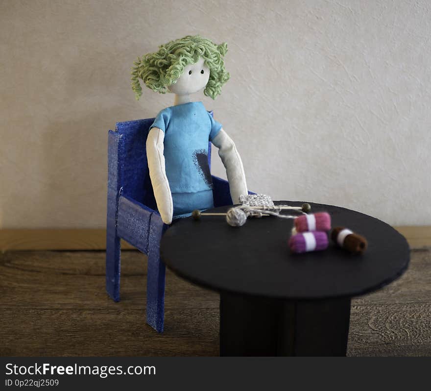 Knitting image. A handmade rag doll is sitting at a black table with a knitting project with wooden knitting needles. Skeins of wool on the table. Room for copy. Knitting image. A handmade rag doll is sitting at a black table with a knitting project with wooden knitting needles. Skeins of wool on the table. Room for copy.