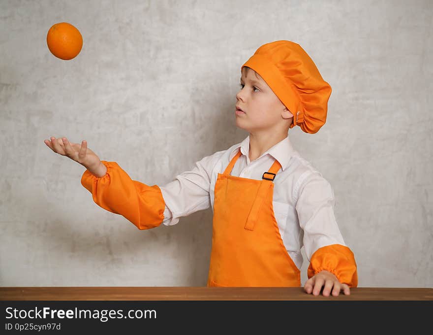 Cute European boy in an orange suit chef tosses an orange up standing at a table on the background of the gray wall. Cute European boy in an orange suit chef tosses an orange up standing at a table on the background of the gray wall