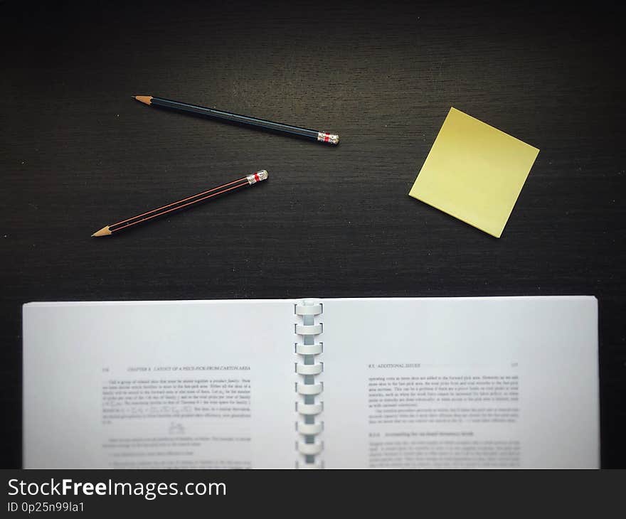 Textbook &#x28;blurred&#x29;, pencils, and yellow sticky note, on the dark wooden table. Textbook &#x28;blurred&#x29;, pencils, and yellow sticky note, on the dark wooden table