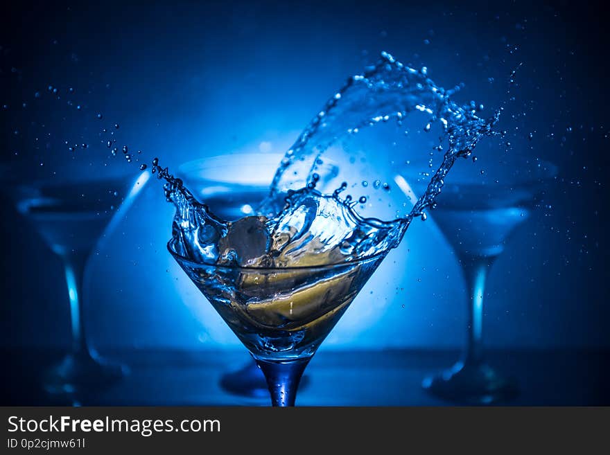 Martini cocktail glass in hand splashing on dark toned smoky background or colorful cocktail in glass with splashes and olives.