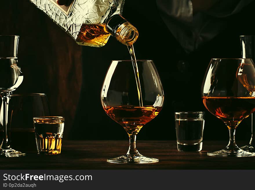 The bartender pours the cognac or brandy in big wine glass on the old bar counter. Vintage wooden background in pub or bar, night