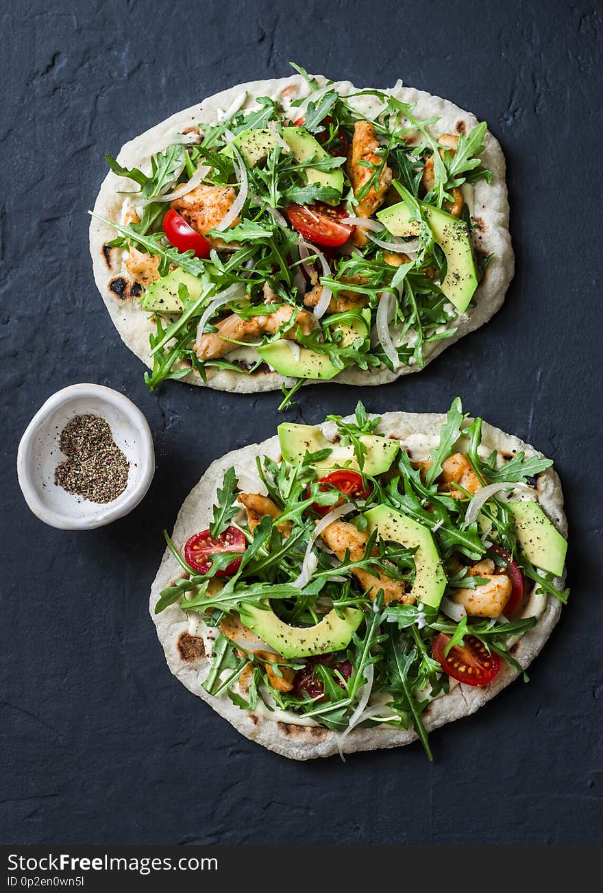 Pan-fried flatbread chicken, avocado, tomatoes, arugula pizza on dark background, top view