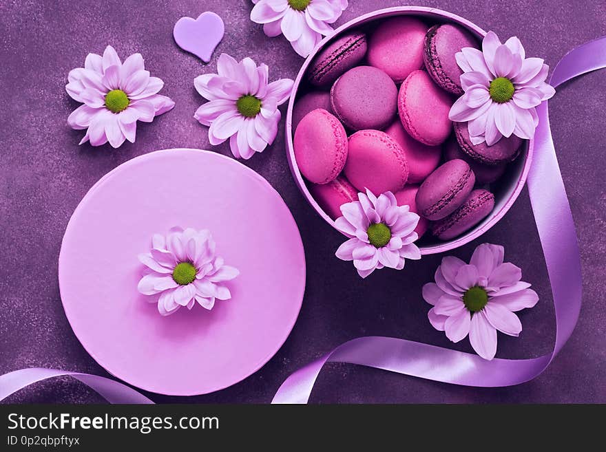 Purple and pink macaroons in a gift box on a beautiful purple background decorated with flowers. Top view.