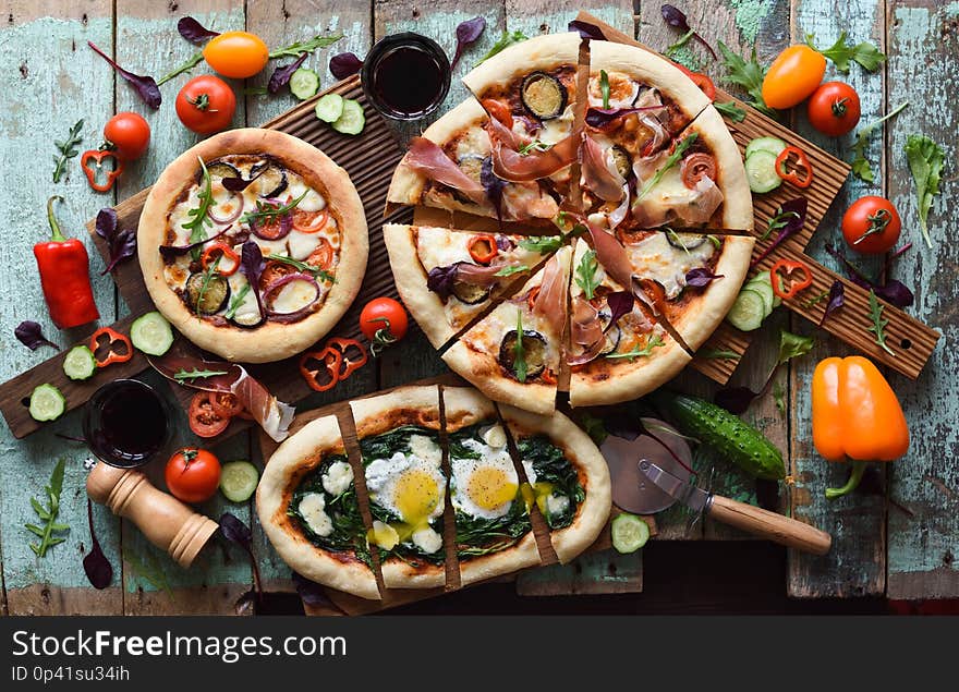 Homemade rustic style pizza party. Flatlay of puffy pizzas with pancetta, aubergines, spinach, eggs, bell pepper and arugula served with raw ingredients on oak boards on shabby blue background overhead view